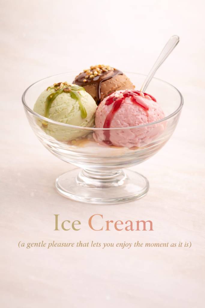 A glass dessert bowl with three scoops of ice cream topped with sauces and nuts, with a spoon placed inside, against a soft light background.